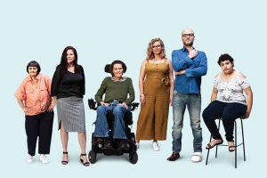 The image shows six people. They have visible and invisible disability. One person is in a wheelchair. One person has a leg brace on their left foot. One person is sitting on a chair. They are positioned in a line.