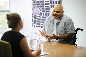 Picture of a man in a wheelchair talking to a woman across a table. 